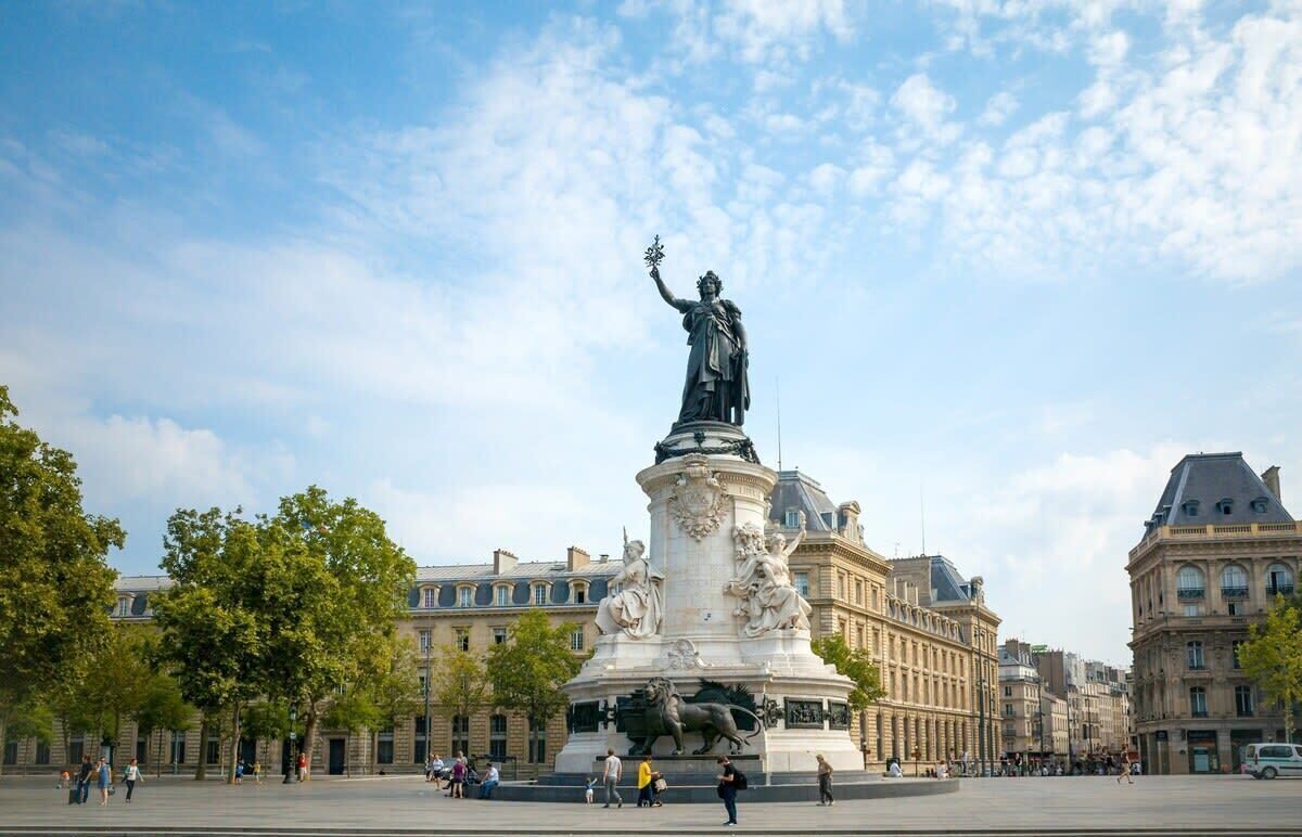 Une vaste place ouverte, avec un monument central sur lequel se trouve une statue d’une femme tenant une torche.