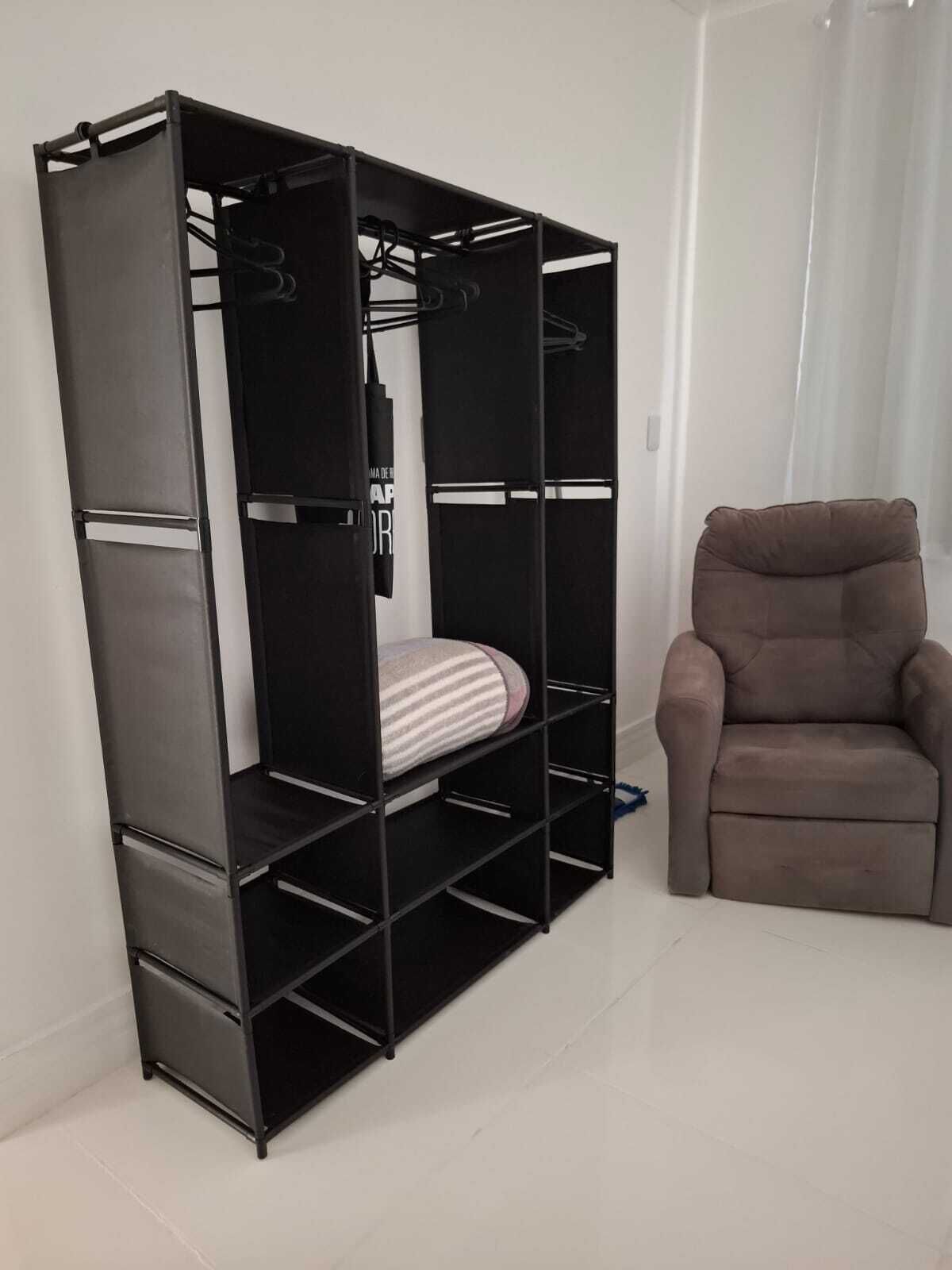 A black wardrobe with multiple shelves and hanging space, a striped cushion, and a brown armchair in a room with white walls and a tiled floor.