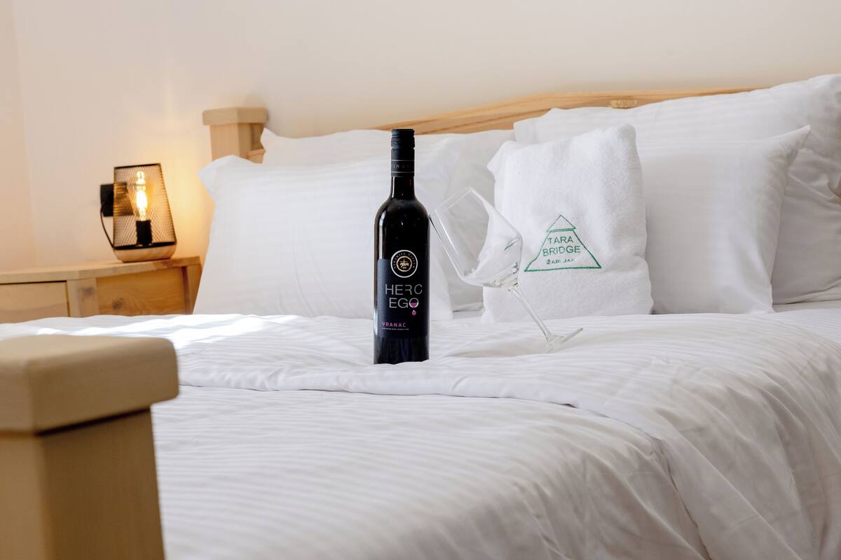 Un lit avec du linge de lit blanc, une bouteille de vin « Herc EGO », un verre et une table de chevet avec une lampe.