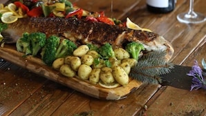 Una mesa de madera con un pescado asado, verduras y una botella de vino.