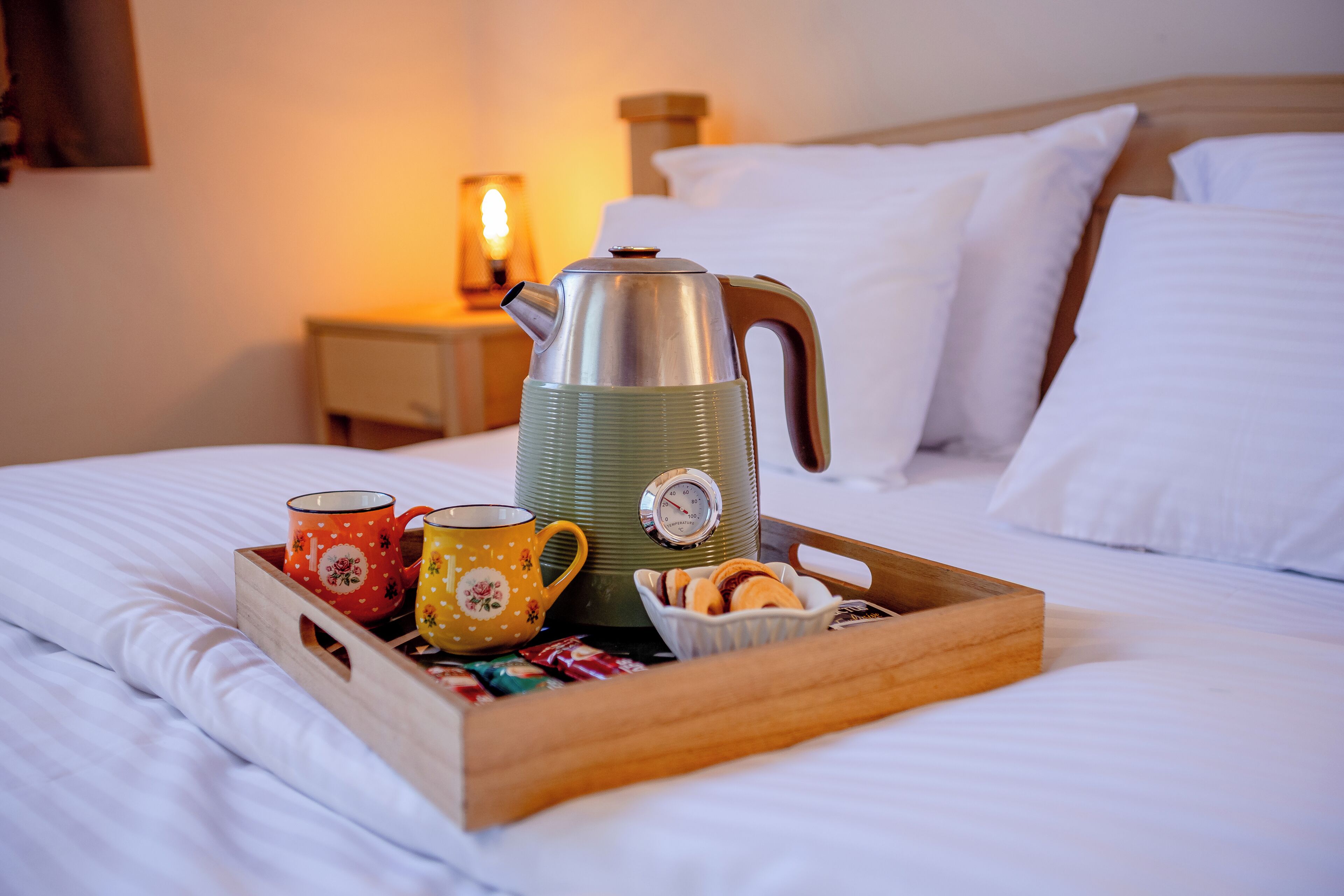 Un plateau en bois avec une bouilloire, deux tasses et un bol de viennoiseries, le tout posé sur un lit.