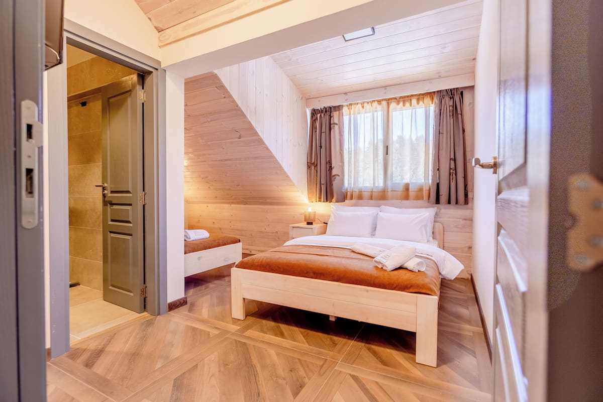 Une chambre avec un plafond en pente en bois, un lit, une table de chevet et une fenêtre avec des rideaux.