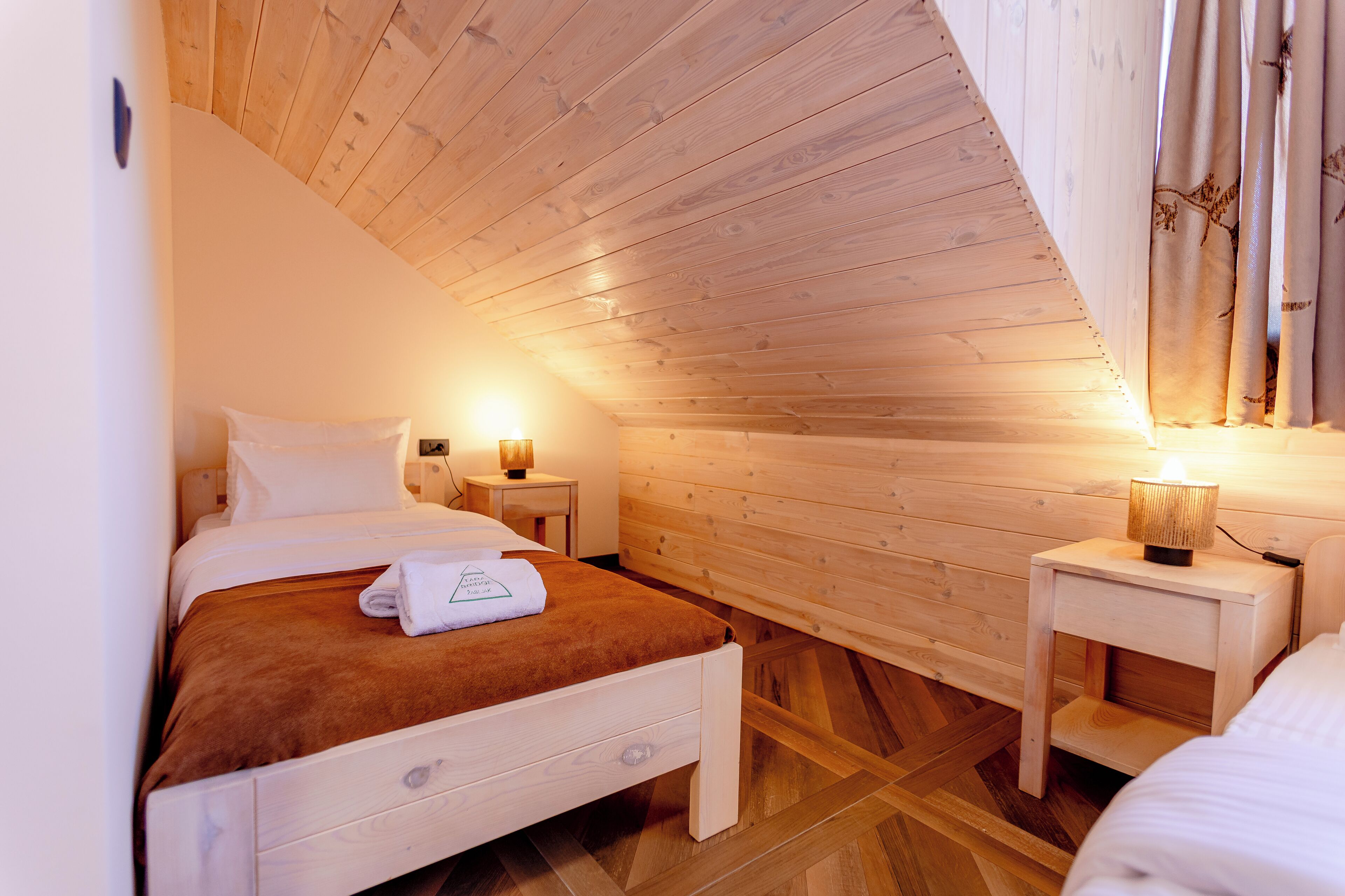 Een slaapkamer met een houten plafond, een bed met een bruine deken, twee nachtkastjes met lampen en een houten vloer.