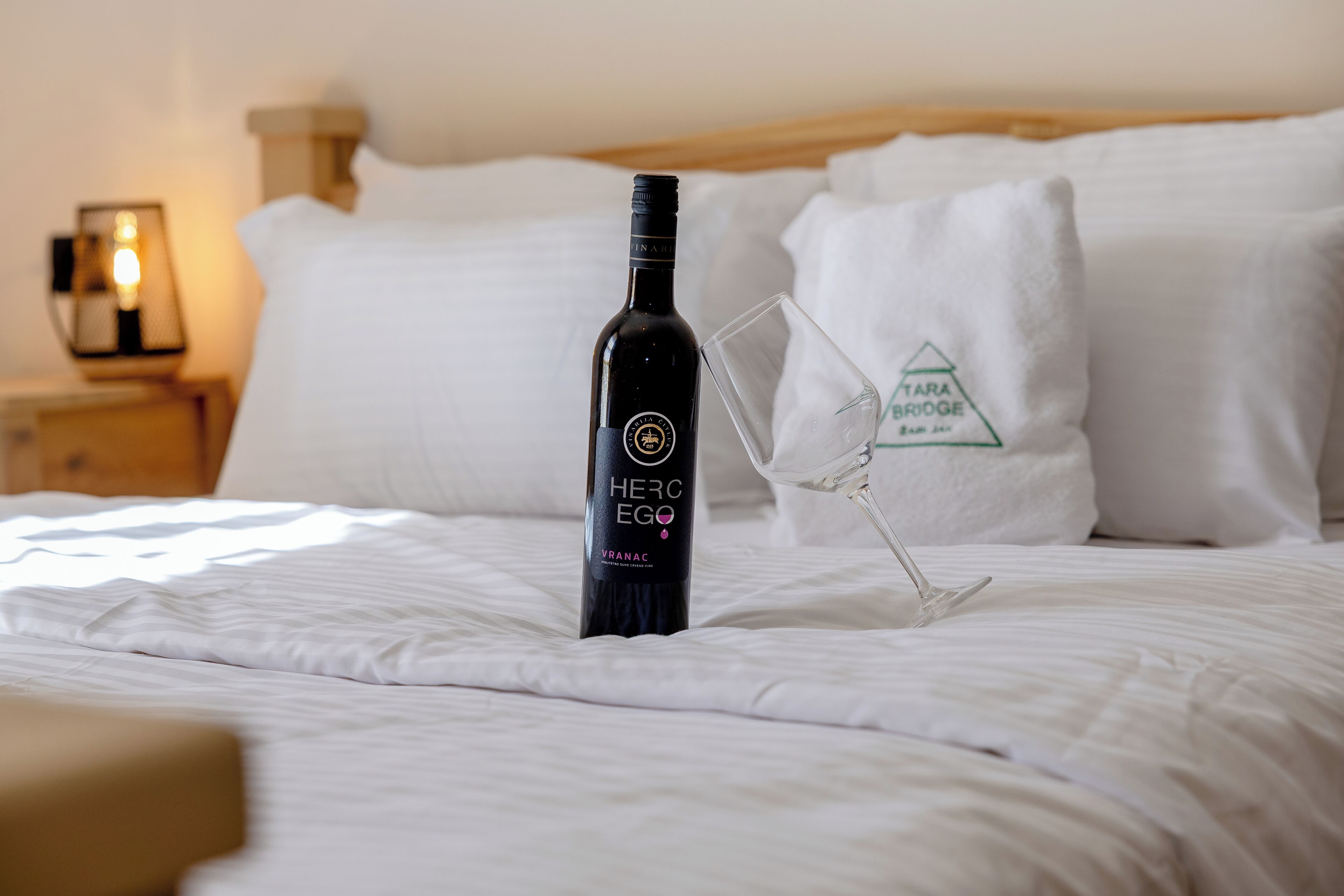 A bottle of wine and a wine glass on a bed with white linens and pillows.