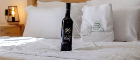Una botella de vino y una copa sobre una cama con sábanas y almohadas blancas.