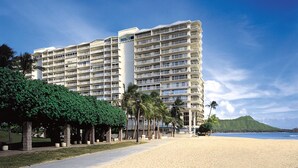 Ein mehrstöckiges Hotel mit Balkonen, im Hintergrund ein Strand und ein Berg.