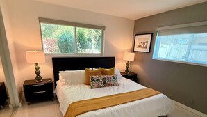 A bedroom with a large bed, two bedside tables with lamps, a window with a view of greenery, and a framed picture on the wall. - Remodeled Mid-century home with pool, hot tub & EV charger (Wilton Manors)