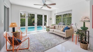 A spacious living area with a sofa, a chair, a ceiling fan, and a view of a swimming pool. - Remodeled Mid-century home with pool, hot tub & EV charger (Wilton Manors)