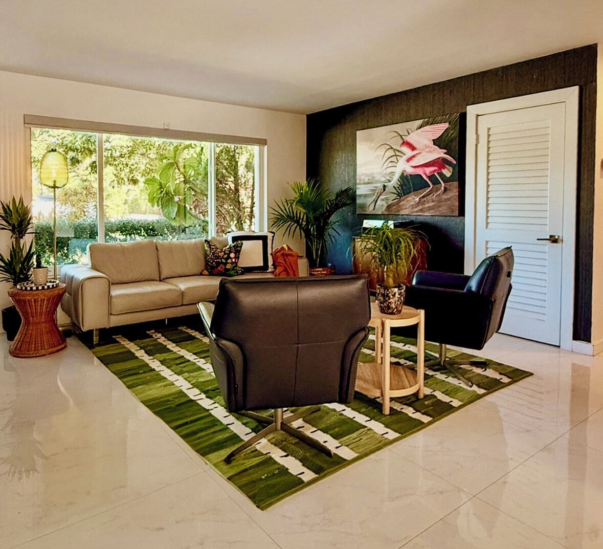 Un salon moderne avec un canapé, un fauteuil et une table d’appoint, de grandes fenêtres et une porte ornée d’un tableau.