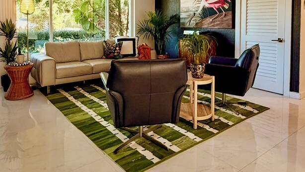 A modern living room with a sofa, armchair, and side table, large windows, and a door with a painting. - Remodeled Mid-century home with pool, hot tub & EV charger (Wilton Manors)