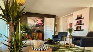A living room with a sofa, a coffee table, a potted plant, a lamp, and a painting on the wall. - Remodeled Mid-century home with pool, hot tub & EV charger (Wilton Manors)