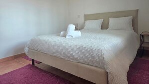 A neatly made bed with a beige headboard and white bedding, a purple rug, and a small bedside table. - Elena's corner (Funchal)