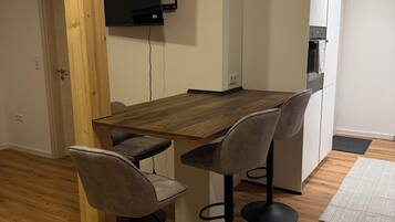 Ein Essbereich mit einem Holztisch und vier Stühlen, einem an der Wand befestigten Fernseher und einer Küchenzeile mit Schränken und einem Backofen.