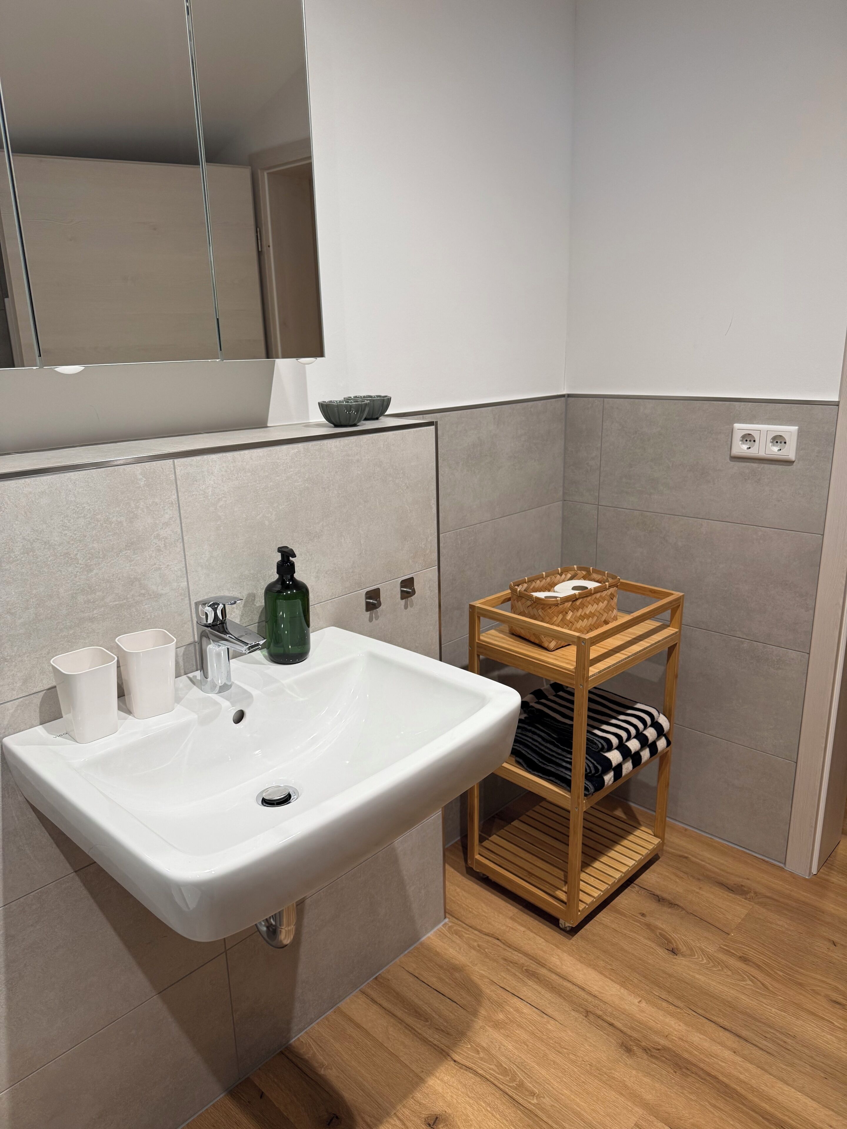 Une salle de bain avec un lavabo blanc, deux tasses blanches, une bouteille verte, une étagère en osier avec des serviettes et un miroir au-dessus du lavabo.