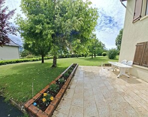 Eine Terrasse mit Tisch und Stühlen, ein Ziegelpflanzgefäß mit Blumen und ein großer Baum.