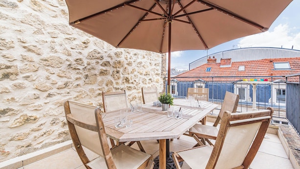 A wooden table with chairs and an umbrella, set against a stone wall with a view of buildings. - Unusual bubbles (Reims)