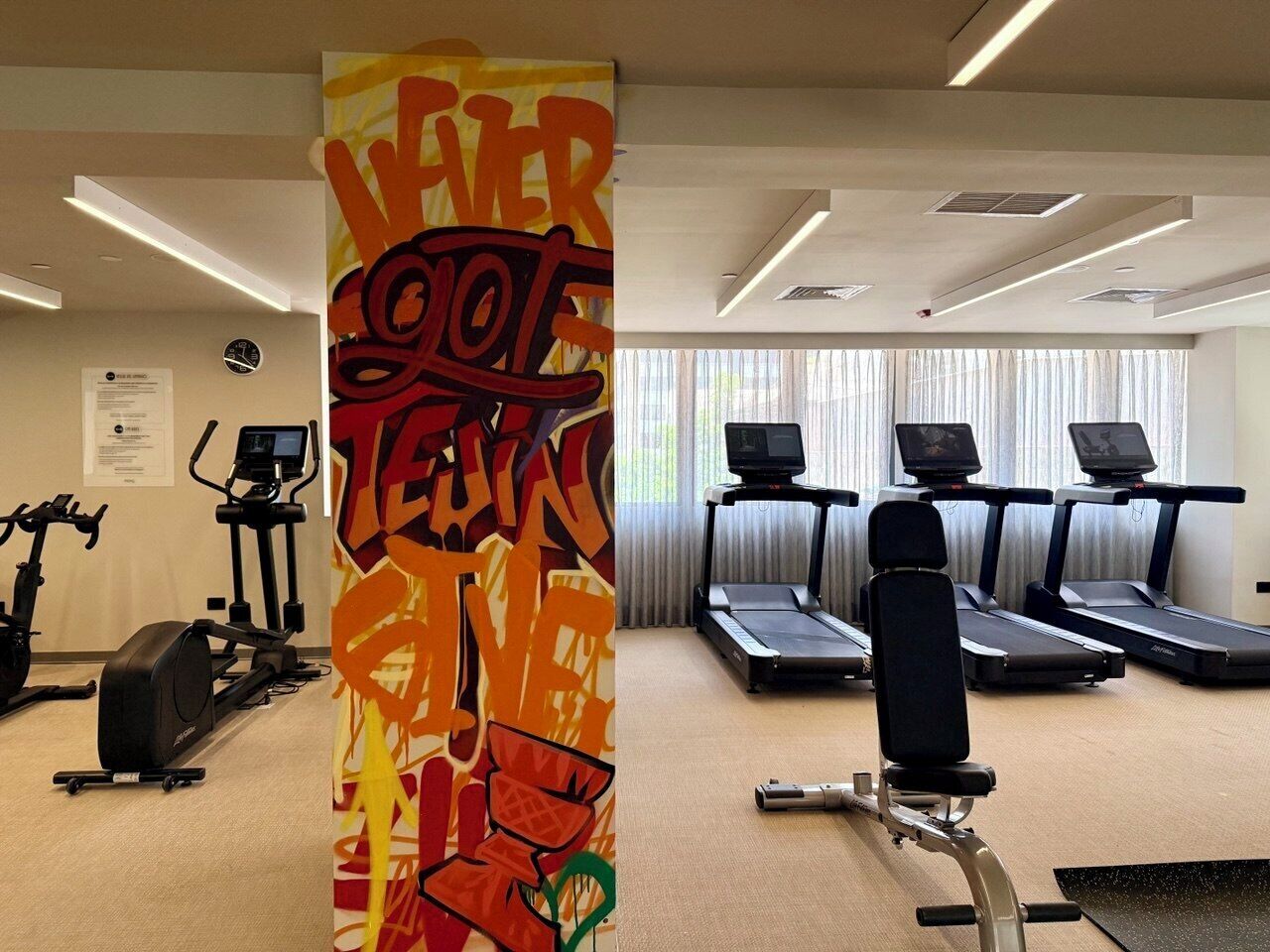 Un gimnasio con cintas de correr, bicicletas estáticas y una pared con un diseño abstracto y colorido.