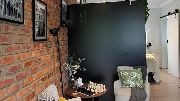 Una sala de estar moderna con pared de ladrillo, pared de acento negra y piso de madera. Hay dos sillones, una mesita de centro y una planta en maceta.