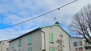 Un edificio de dos pisos con contraventanas verdes, una chimenea visible y un pequeño árbol en la parte delantera.