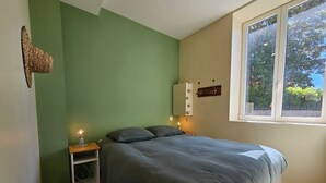 Ein Schlafzimmer mit Bett, Nachttisch mit Lampe, Wandregal und Fenster mit Blick auf Bäume.