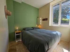 Un dormitorio con cama, mesita de noche con lámpara, estante en la pared y ventana con vista a árboles.