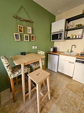Cocina compacta con zona de comedor, que incluye una mesa y sillas de madera, un microondas, un fregadero y un estante pequeño con utensilios de cocina.
