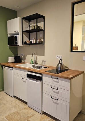 Cocina pequeña con armarios blancos, encimera de madera, microondas integrado, hervidor y un estante con cristalería.