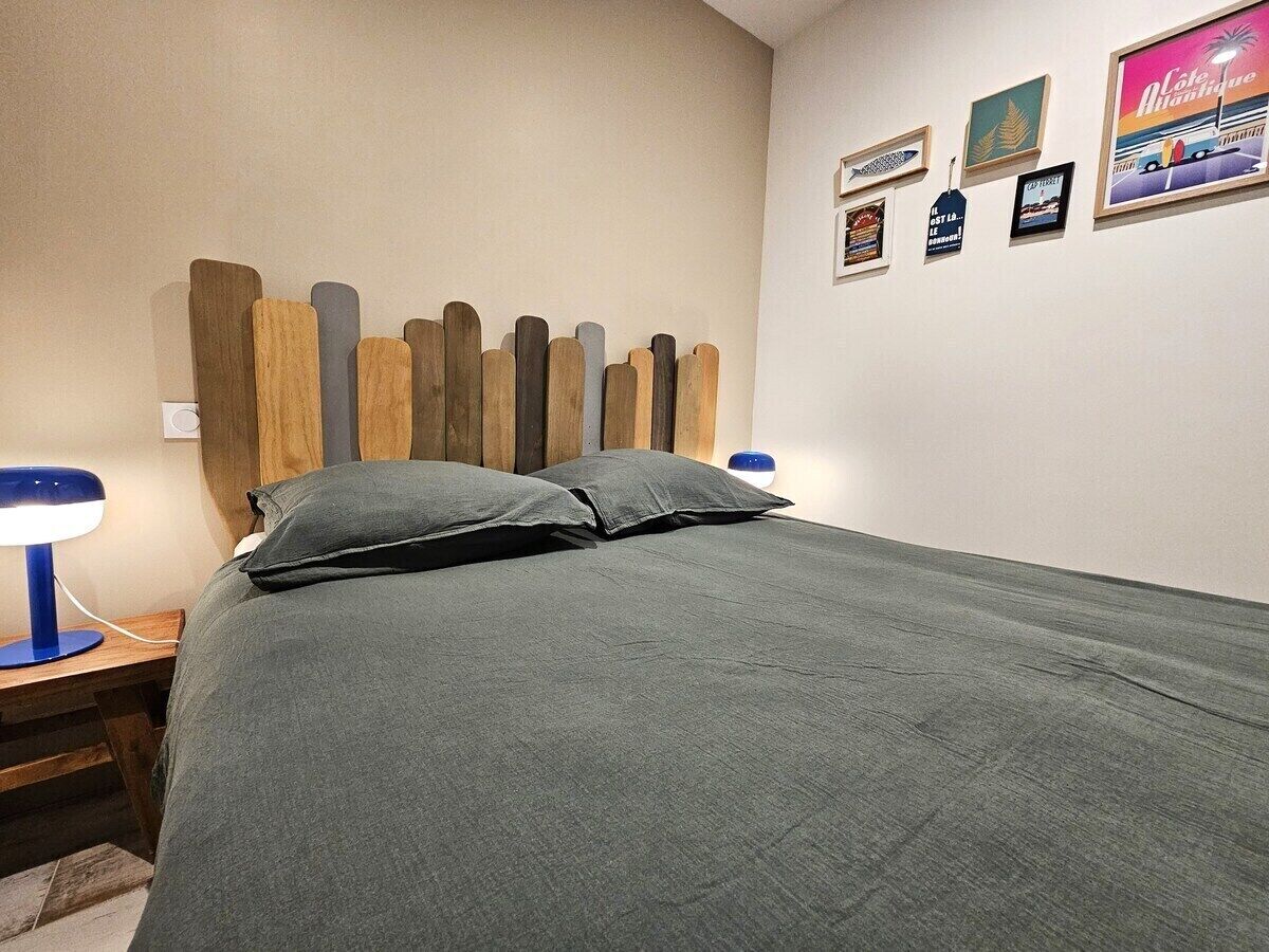 Een net opgemaakt bed met een grijze spreidek en kussens, een houten hoofdbord, en een nachtkastje met een blauwe lamp.