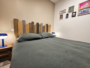 Ein ordentlich bezogenes Bett mit einer grauen Tagesdecke und Kissen, ein Holz-Kopfteil und ein Nachttisch mit einer blauen Lampe.