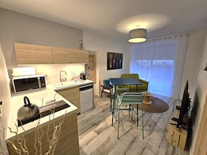 Et moderne køkken med mikrobølgeovn, elkedel og vask, et spiseområde med bord og stole, samt et stort vindue med gardiner.