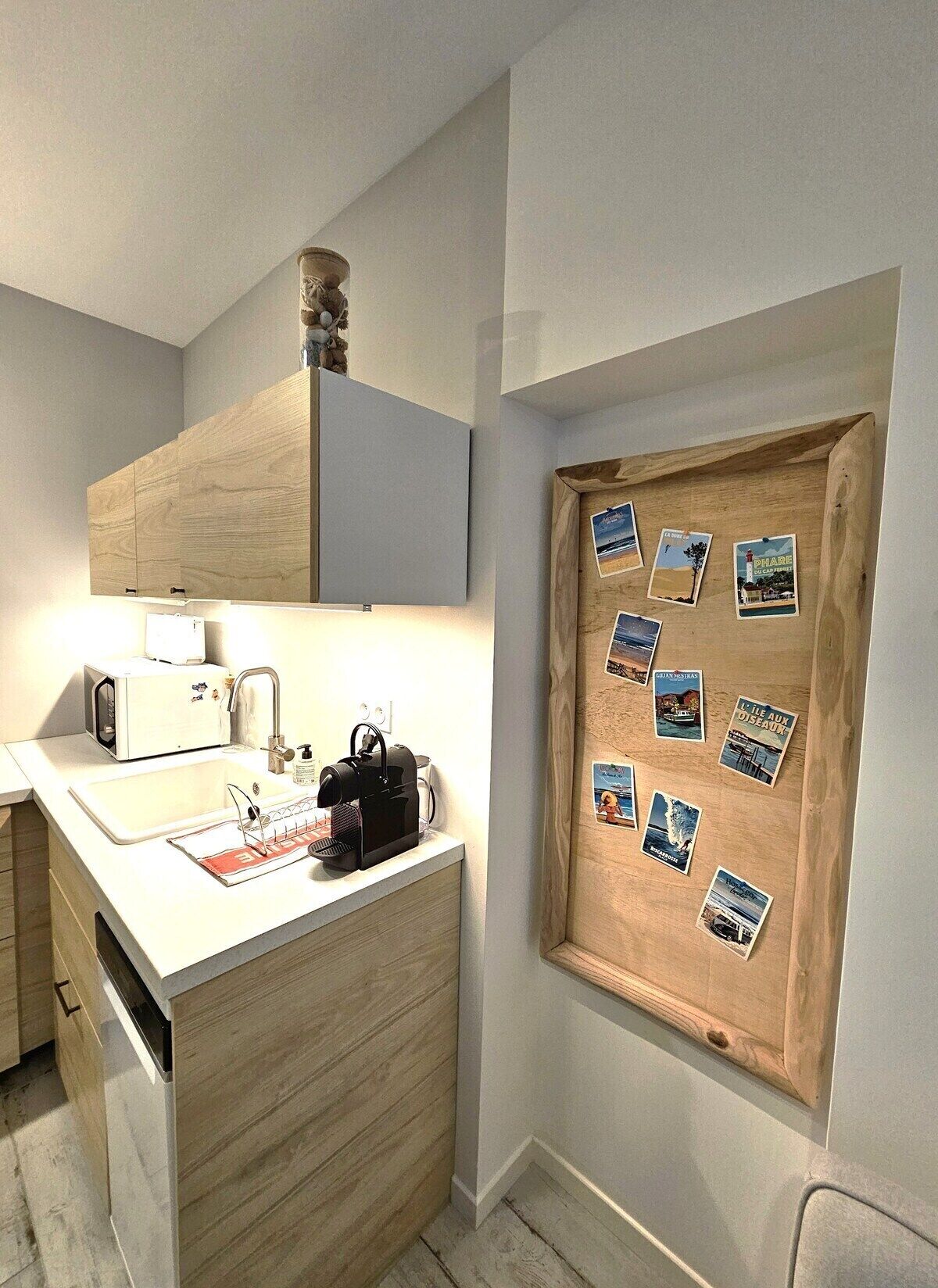 Une cuisine compacte équipée d’un meuble en bois, d’un four à micro-ondes, d’une machine à café et d’un tableau d’affichage en liège où sont accrochées des photos de voyages.
