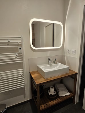 Une salle de bain moderne avec un lavabo blanc, un miroir avec éclairage intégré, un porte-serviettes et une étagère en bois où sont rangés les articles de toilette.