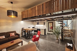 Un salon moderne avec un canapé, une table basse et un fauteuil rouge. On y trouve également un coin cuisine avec une table à manger et un escalier menant à l’étage supérieur.