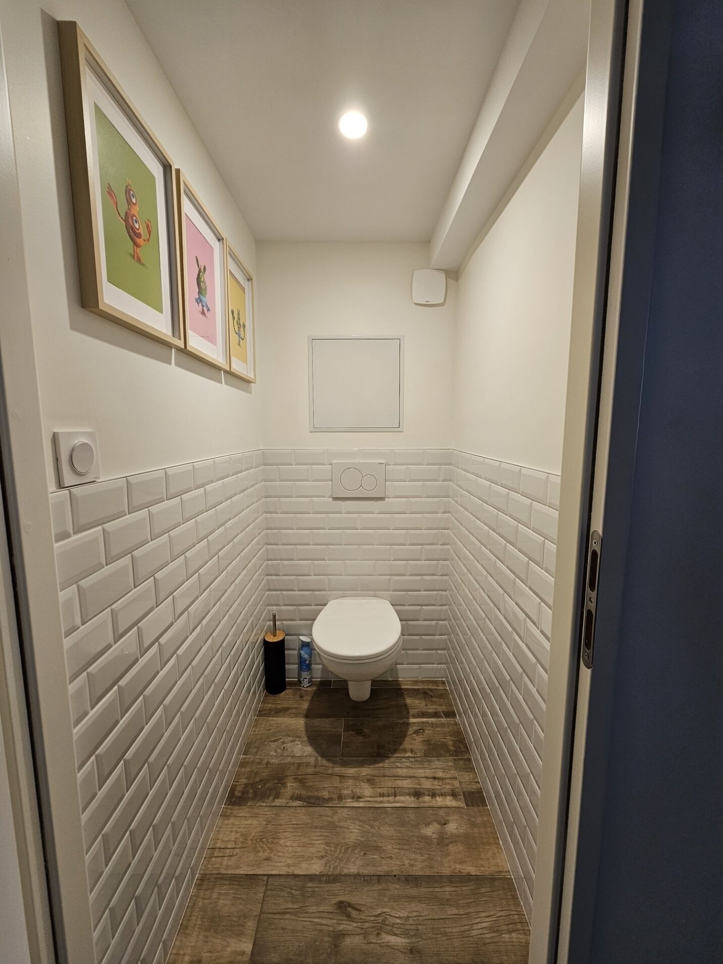 Een smalle badkamer met witte betegeling, een wit toilet en een houten vloer. Er hangen drie afbeeldingen in lijstjes aan de muur.