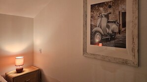 Une photo ancienne d’un scooter encadrée, accrochée au mur, une lampe de chevet et une table de nuit en bois.
