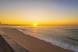 Praia com pôr do sol, ondas e uma barreira de concreto.