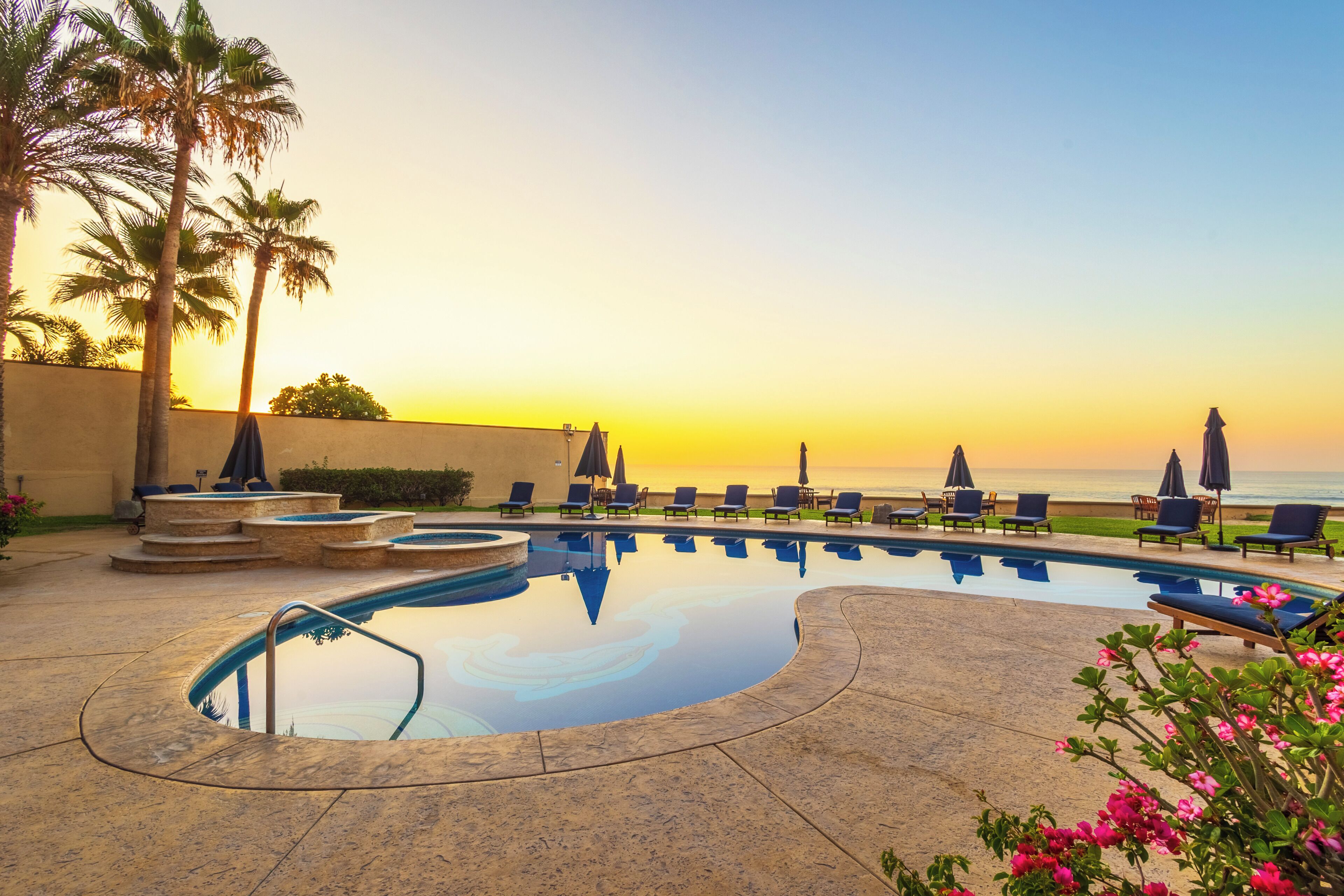 A pool area with lounge chairs and umbrellas, palm trees, and a view of the ocean at sunset.