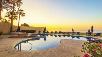 A pool area with lounge chairs and umbrellas, palm trees, and a view of the ocean at sunset.