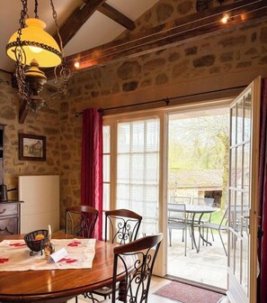 A dining area with a stone wall, a chandelier, a table with chairs, and a view of the outdoors through large windows. - Gîtes de France® - Domaine de Yère (FOURMAGNAC)