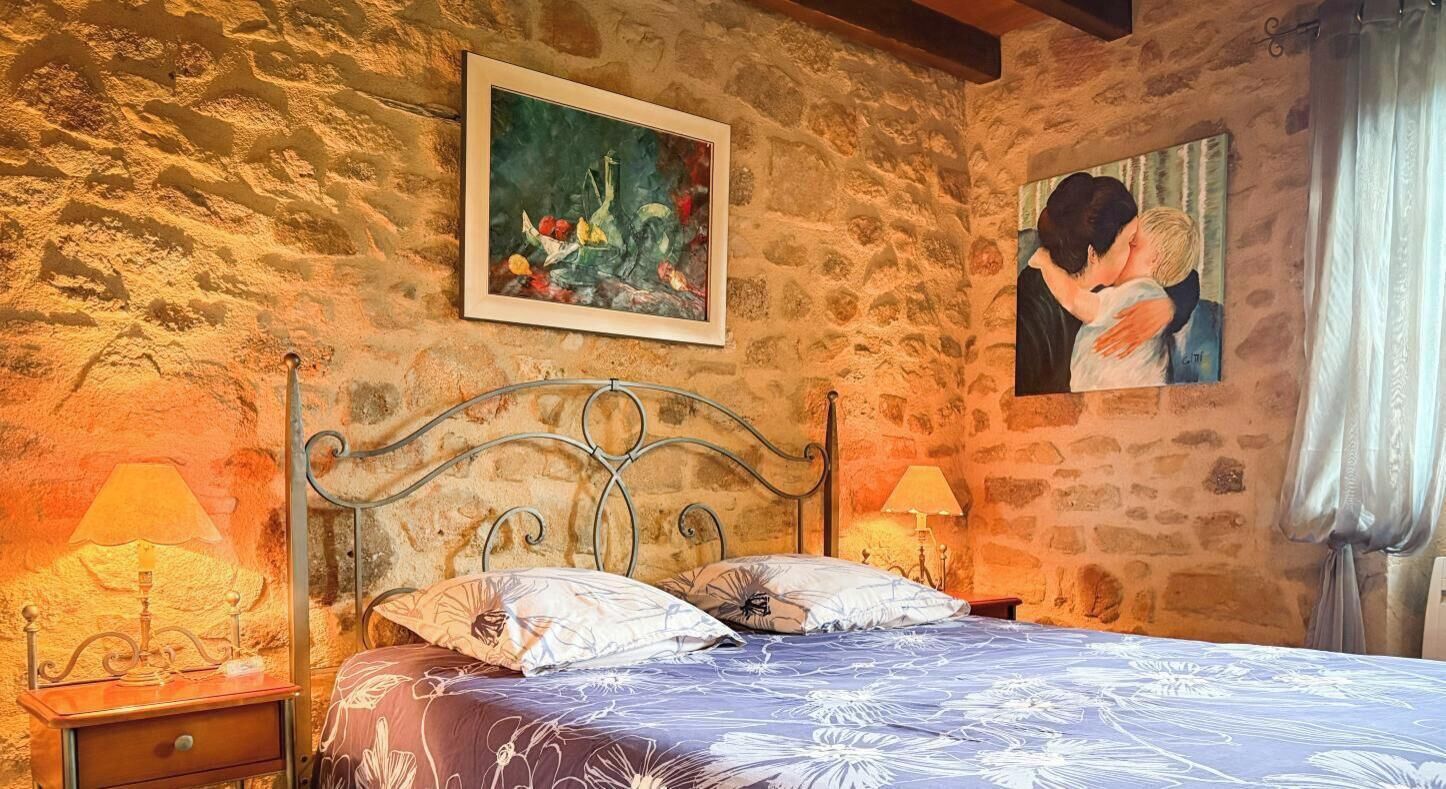 A bedroom with a stone wall, a metal bed frame, two bedside tables with lamps, and two paintings.