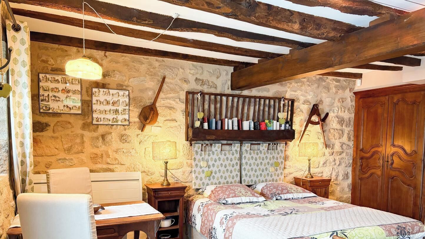 A rustic bedroom with exposed wooden beams, a stone wall, a bed with patterned bedding, bedside tables with lamps, and a wooden wardrobe.