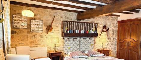 A rustic bedroom with exposed wooden beams, a stone wall, a bed with patterned bedding, bedside tables with lamps, and a wooden wardrobe.