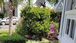 Un jardín bien cuidado con arbustos variados y una palmera, un coche aparcado y una silla rosa cerca de un edificio.