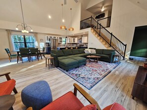 A spacious living area with a sectional sofa, ottomans, a coffee table, and a dining set. A staircase with a railing leads to an upper level.