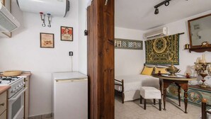 Eine kompakte Küche mit Herd, Kühlschrank und Wand-Klimaanlage. Im Hintergrund ist ein Bett mit Holz Kopfteil und einer dekorativen Wandweberei zu sehen.
