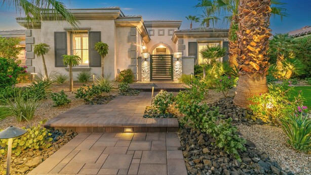 Una casa con facciata in pietra, ingresso con cancello e giardino curato con palme e arbusti.