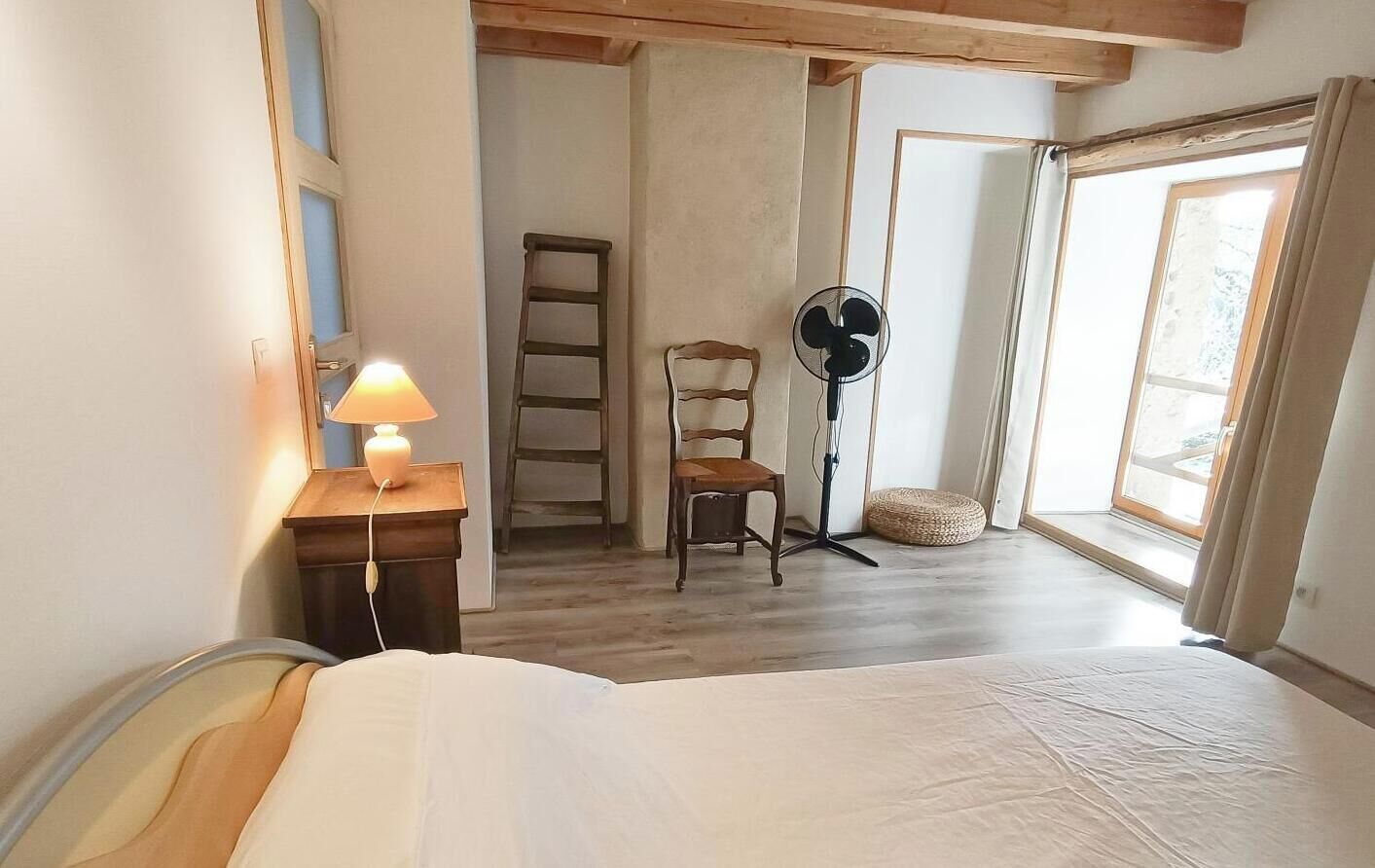 Une chambre à coucher avec un lit, un ventilateur sur pied, une chaise, une échelle et une fenêtre avec des rideaux.