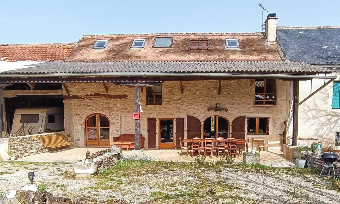A stone building with a tiled roof, a covered patio with wooden furniture, and a barbecue grill.