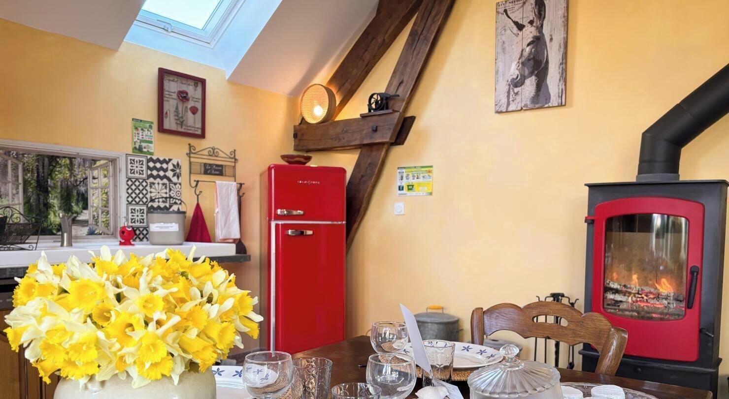 A cozy room with a red stove, a dining table with a vase of flowers, and a window overlooking greenery.