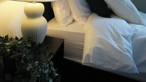 En pænt redt seng med hvide sengetøj, en natlampe og en potteplante på et natbord.